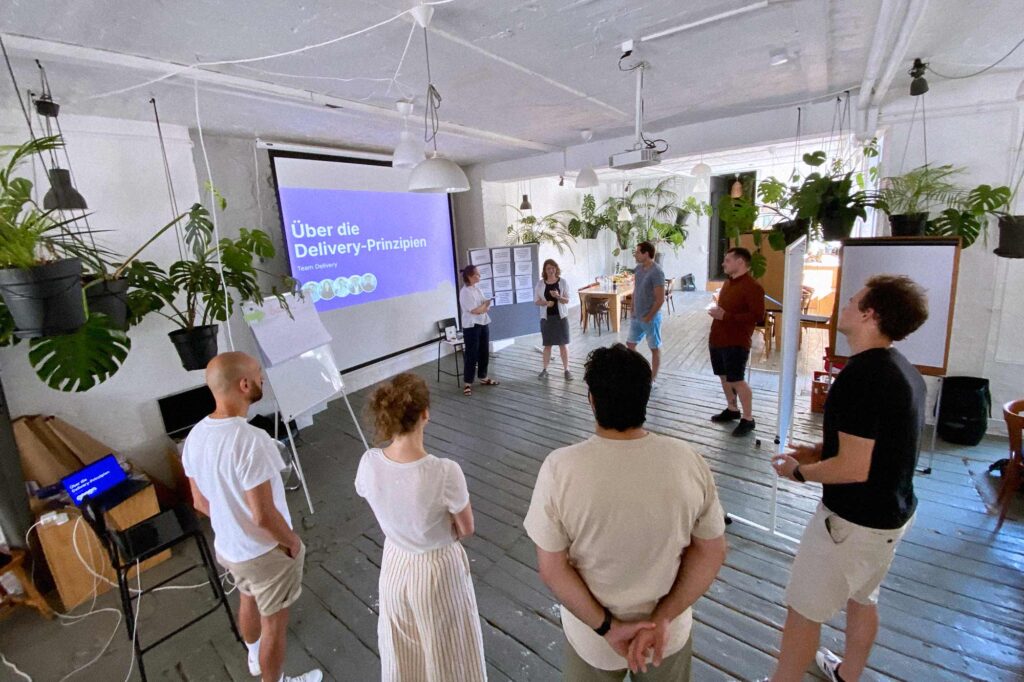 A group of people in casual attire stands in a room with hanging plants, listening to a presentation. A projected slide on the wall reads "About the delivery principles," and there are whiteboards and flip charts nearby. The setting appears to be informal and collaborative.