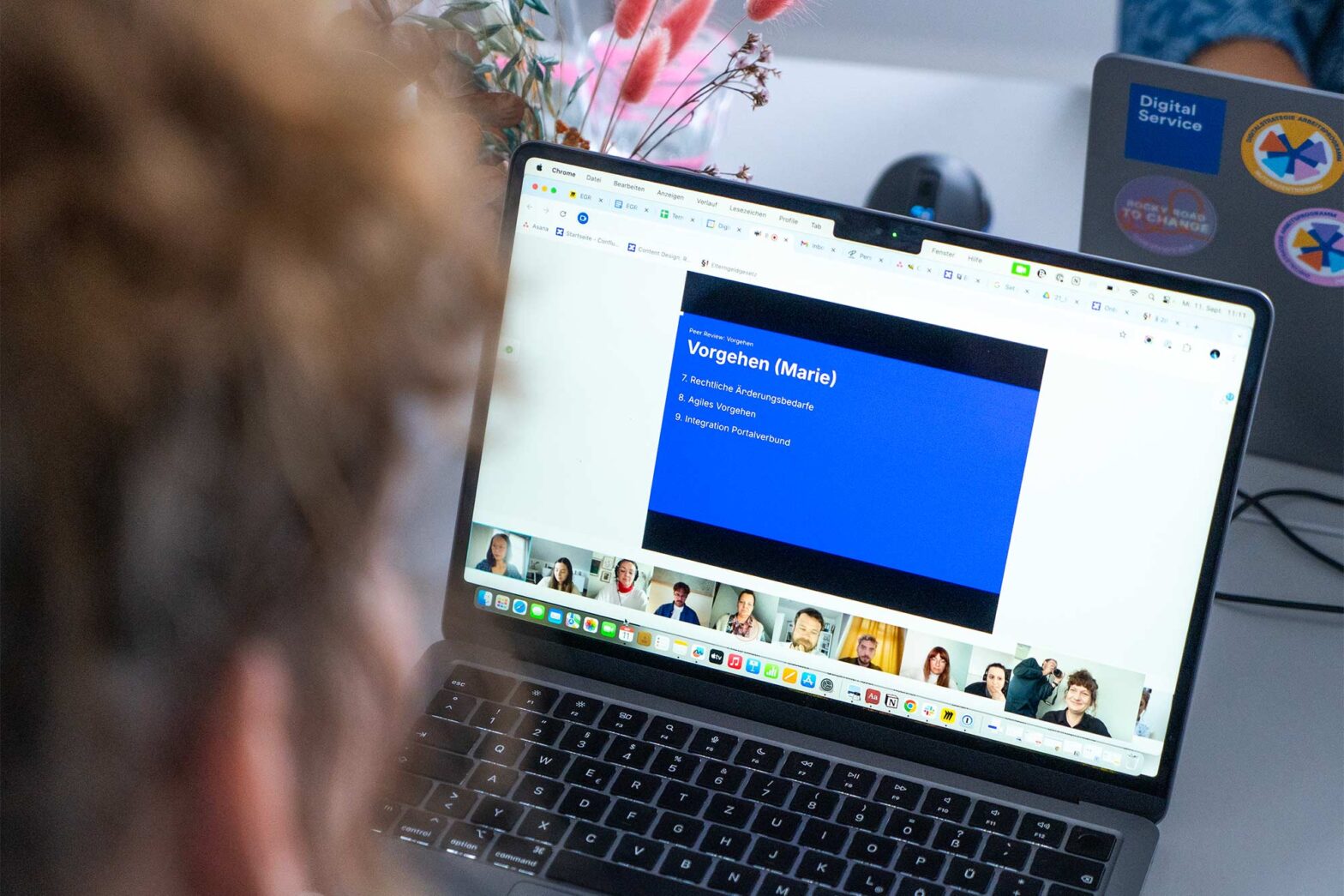 A person is viewing a virtual meeting on a laptop. The screen displays a presentation slide with the title "Vorgehen (Marie)" and a list of bullet points. Below the slide, several participants' video thumbnails are visible in a row. In the background, another person works on a laptop with stickers that read "Digital Service" and "Ask for Help to Change."