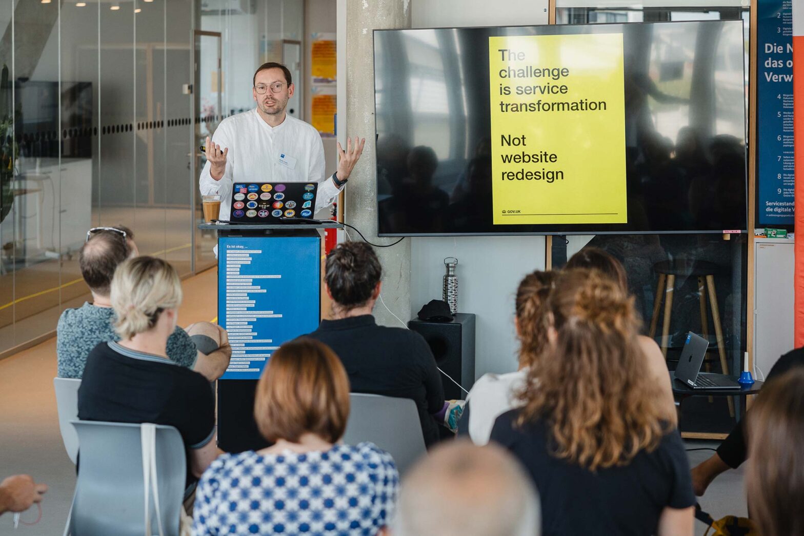 A speaker is presenting to an audience in a modern office setting. The slide on the screen reads, "The challenge is service transformation, not website redesign." The audience is seated and listening attentively, while the presenter gestures with his hands, emphasizing his point. A laptop with multiple stickers is placed on a lectern, and a coffee cup is next to the laptop.