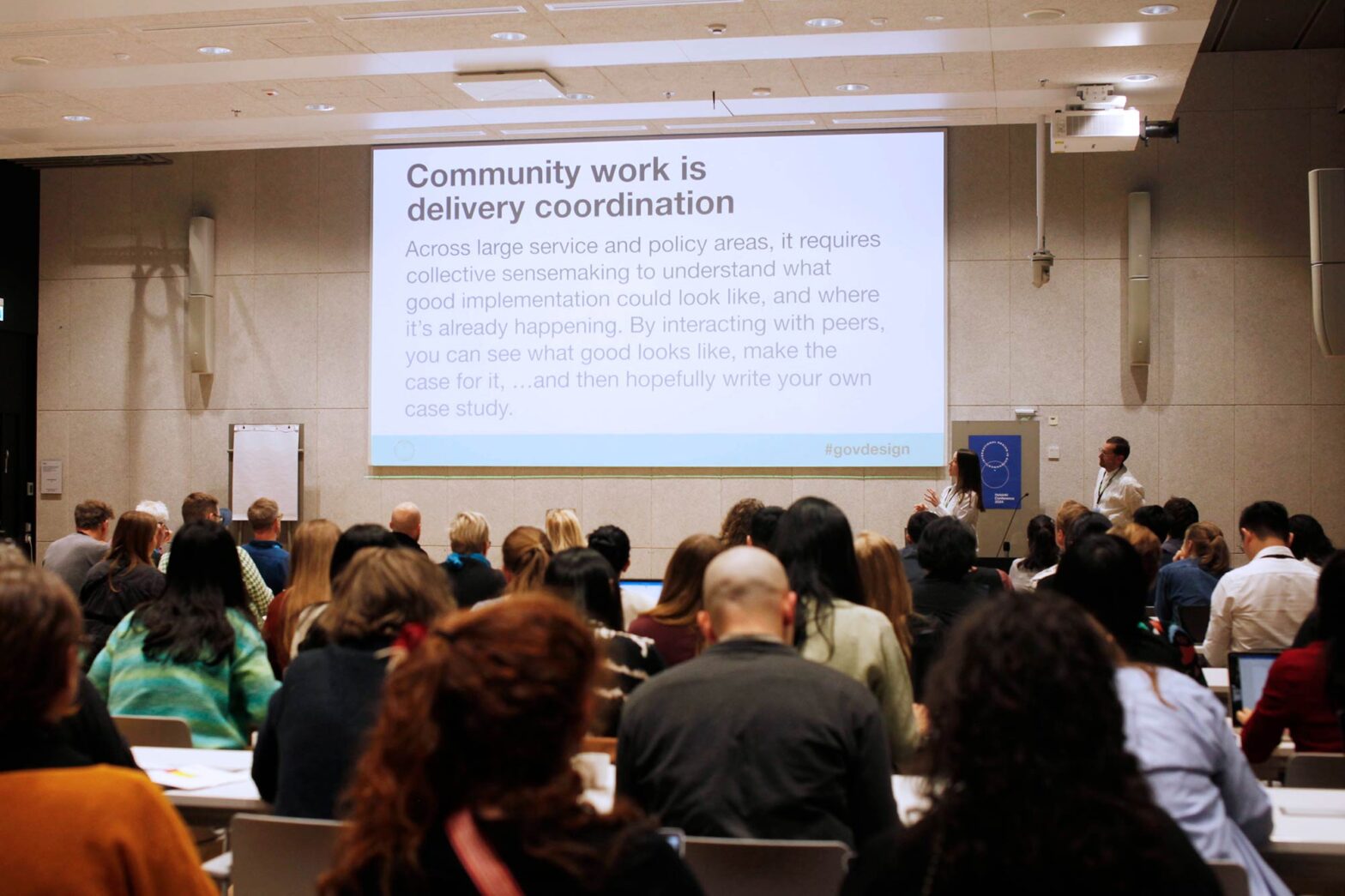 Two speakers are presenting at a conference in front of an audience, with a large slide projected that reads, "Community work is delivery coordination." The text on the slide discusses the importance of collective sensemaking and good implementation in large service and policy areas. The room is filled with attentive participants, some taking notes. The atmosphere suggests a formal and educational setting.