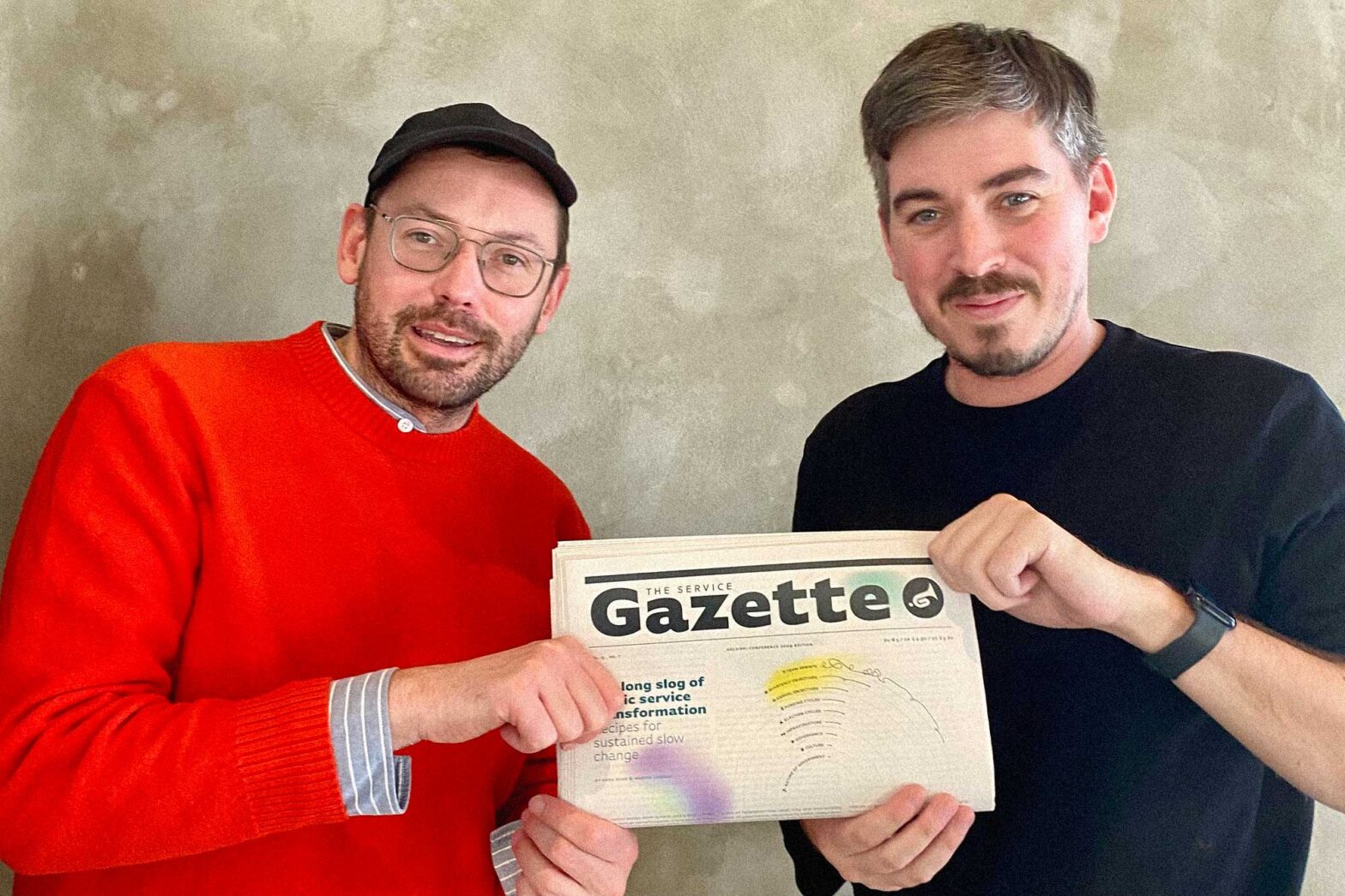 Two men with light skin and in casual clothes standing next to each other, holding a newspaper together. The masthead reads: Gazette, the headline below says: Long slog of service transformation – recipes for sustained slow change