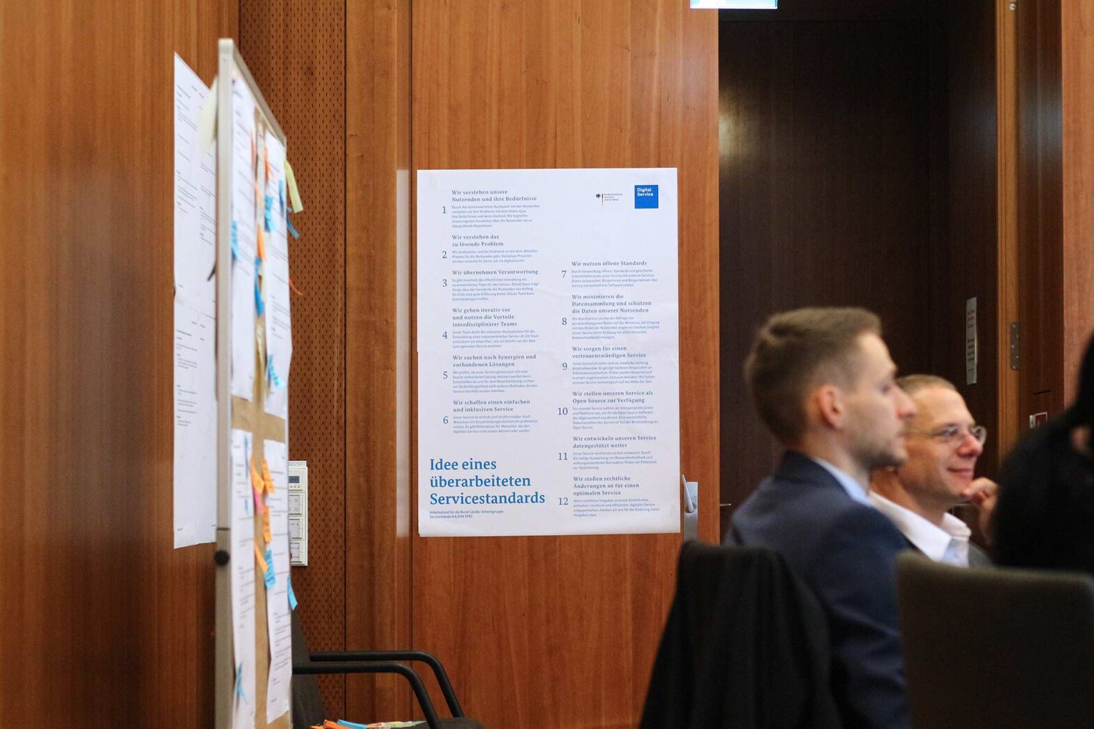 The image shows a workshop setting with people seated in front of a wooden wall. A poster titled "Idee eines überarbeiteten Servicestandards" (Idea of a revised service standard) is visible on the wall, listing twelve points in German. To the left, a board with various notes and sticky notes is partially visible. The focus appears to be on discussions or presentations related to service standards.