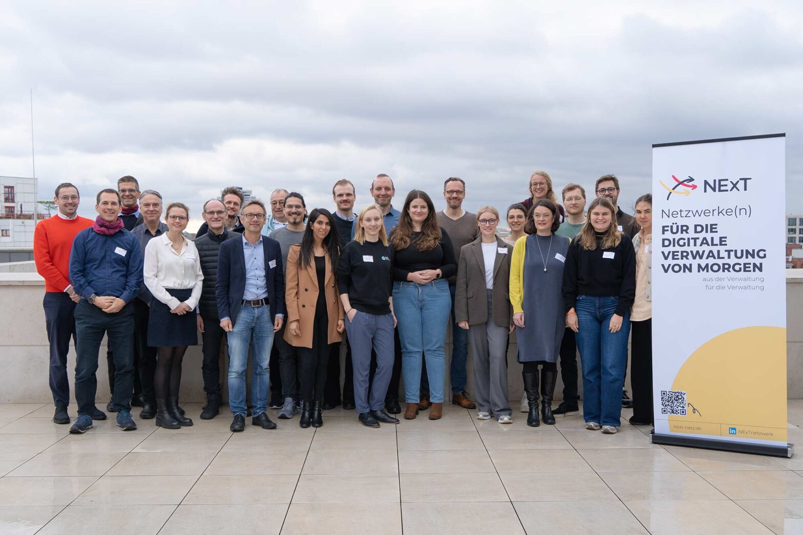 A formal group photo of about 2 dozen people, most of them with light skin, half women, half men – on a roof top with grey clouds above – many people smiling