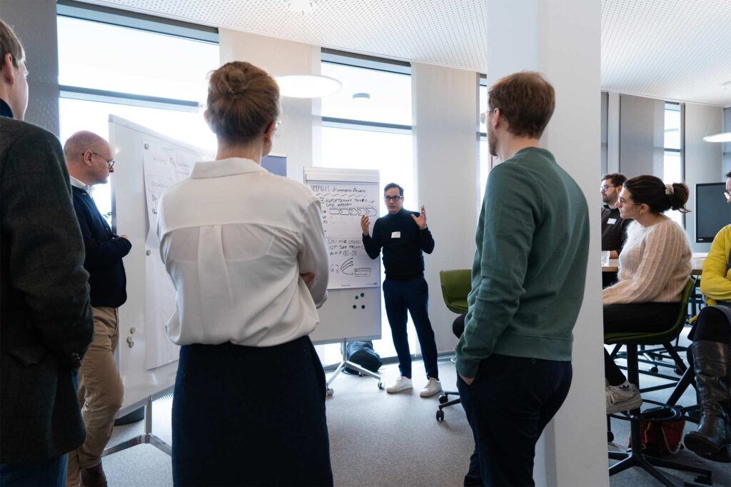 8 people, men and women, in casual business outfits in an open, modern office space with whiteboards and flipchart boards – one man is speaking, gesturing with both hands in front of a process diagram drawn on the board