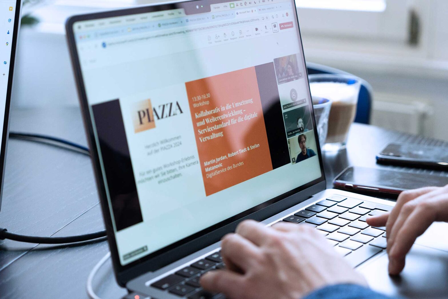 Closeup of a laptop computer showing a screen with the logo of the PIAZZA conference, the talk title: “Collaboratively to implementation: Service Standard for German administration” with 4 small camera views of people next to it