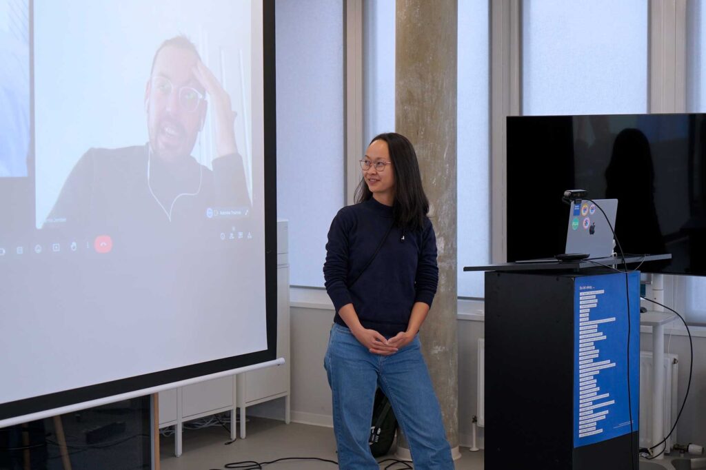 A woman is standing next to a podium with a laptop, smiling and looking at a large screen displaying a video call with a man wearing glasses and headphones. The room is brightly lit with large windows in the background.
