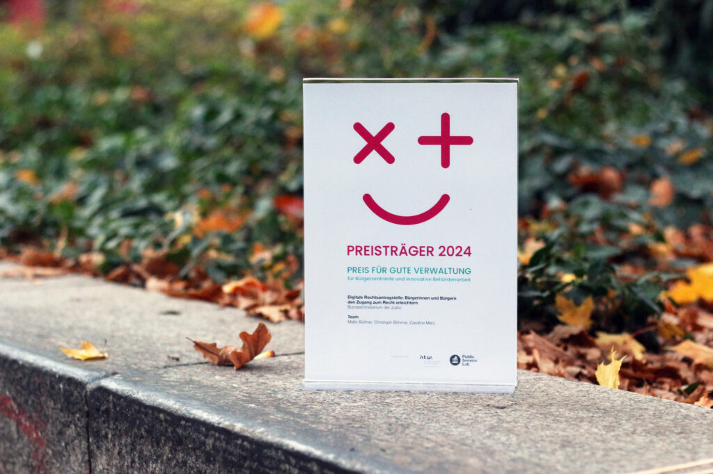 An award certificate with an abstract winking smiley face saying awardee 2024, standing outside on a concrete rim in front of autumn leaves