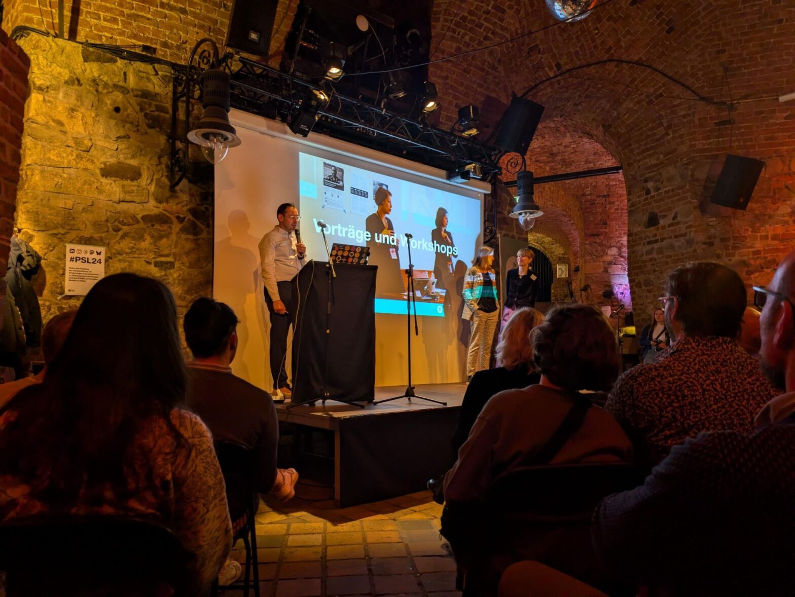 3 people, a man and two women, on small stage in a medieval brick basement with about a dozen people sitting and looking at them