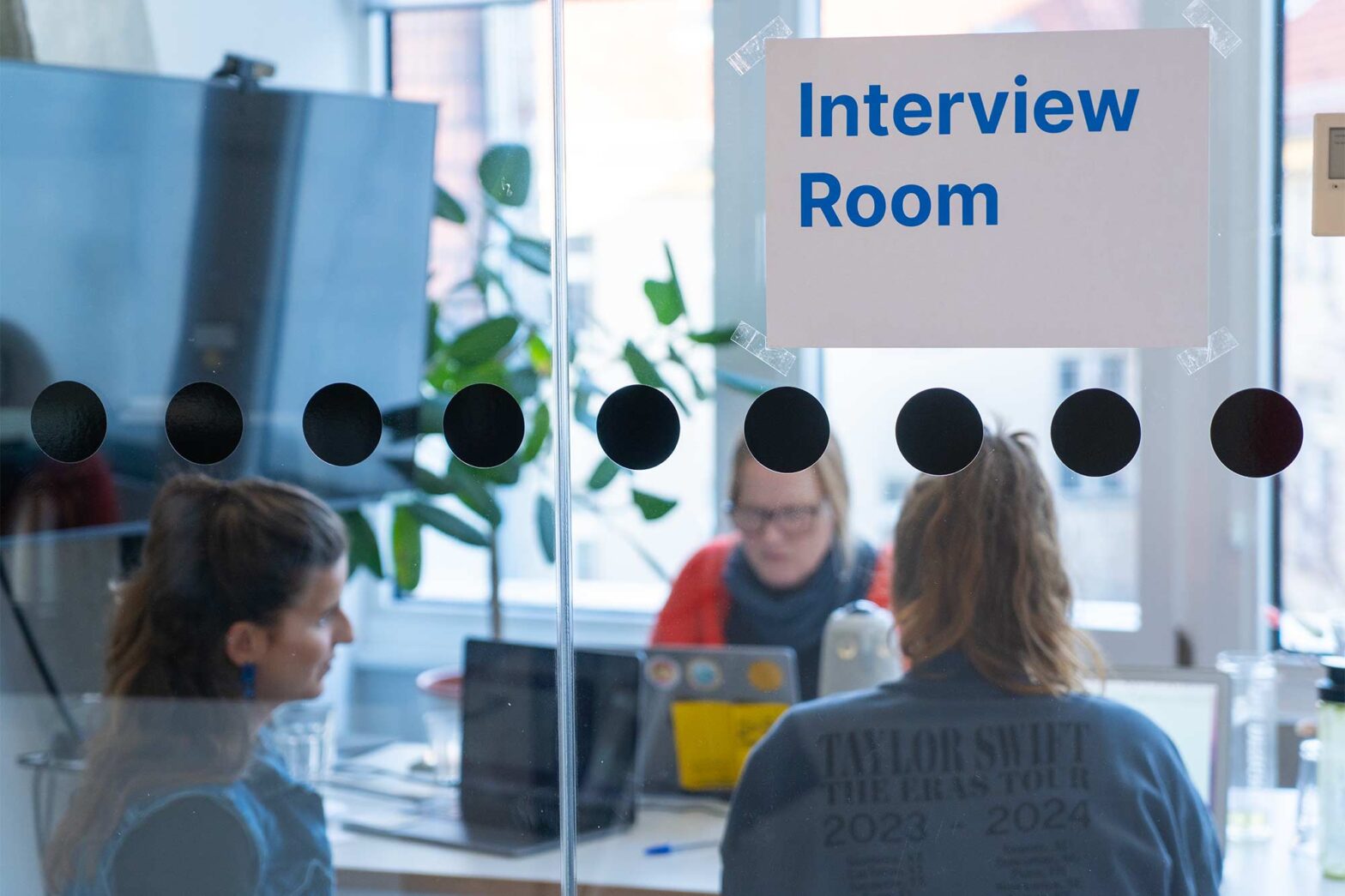 A glass-walled interview room with three people seated inside, two facing away and one facing forward, working on laptops. Sign on glass reads 'Interview Room.'