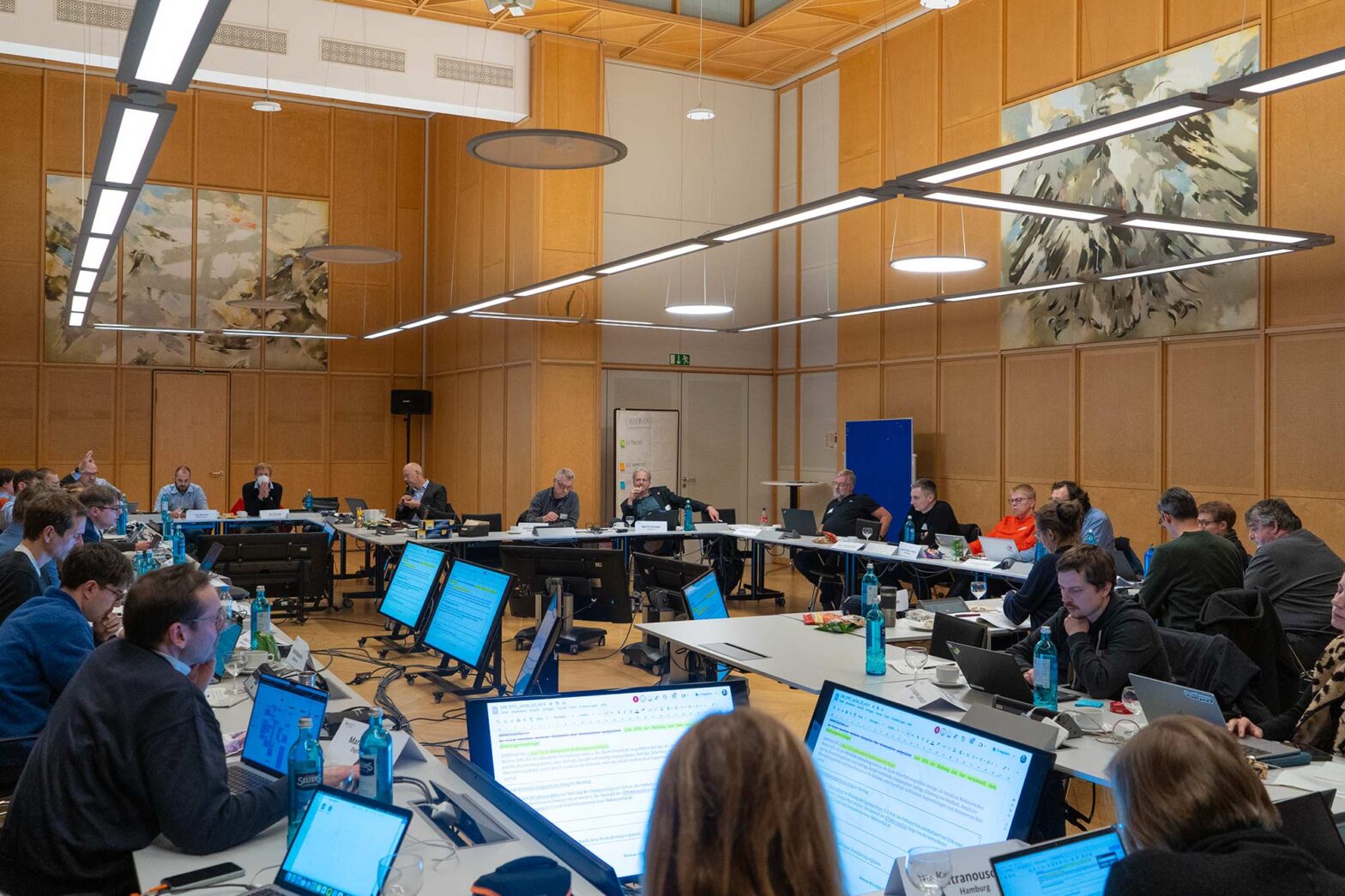 A group of people in a conference room seated at a U-shaped arrangement of tables, working on laptops and documents. Multiple monitors display text, and the room features wooden paneling with large abstract paintings on the walls.