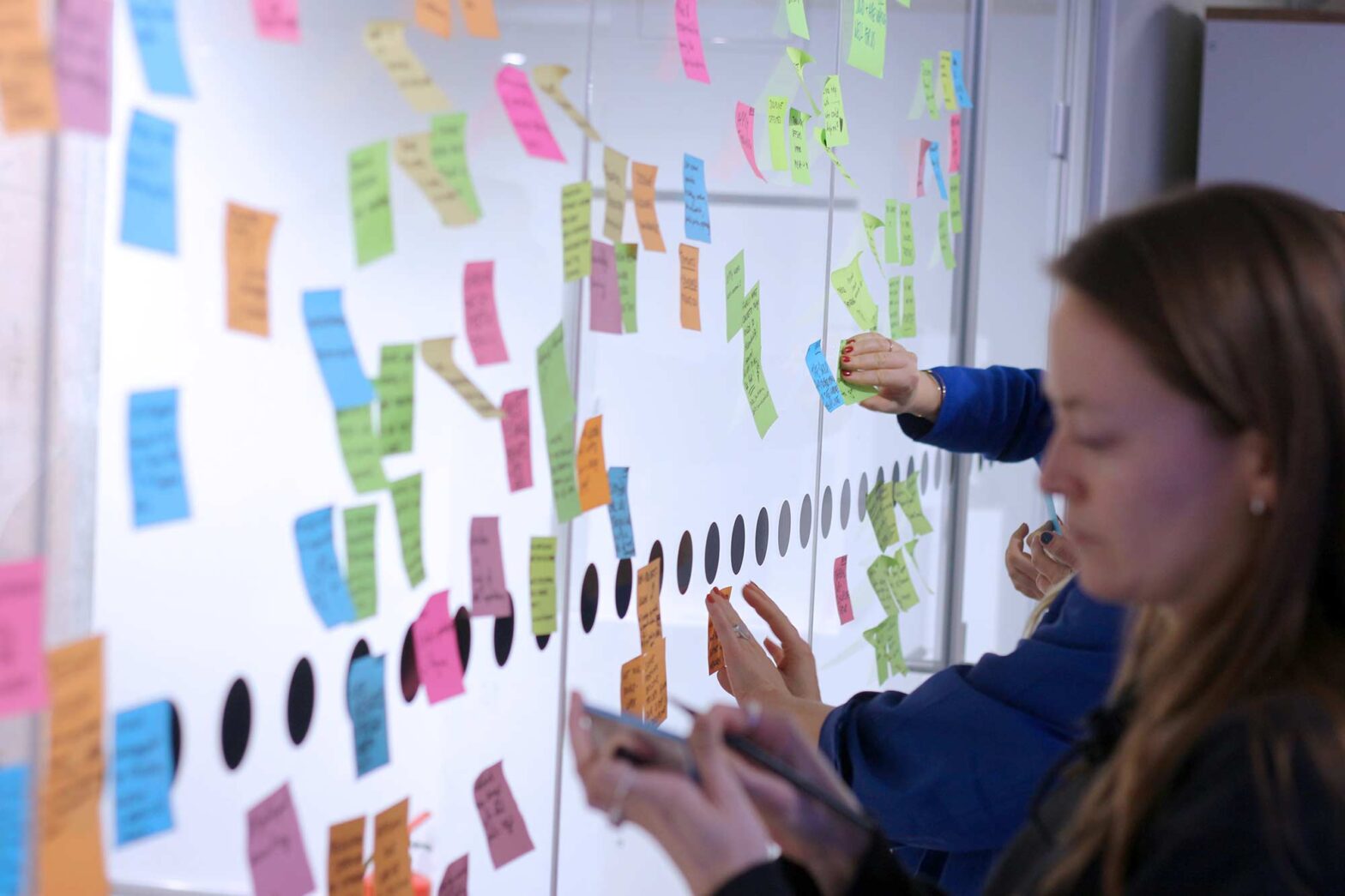 A group of people is brainstorming in front of a whiteboard covered in colorful sticky notes, writing ideas and organizing them collaboratively.