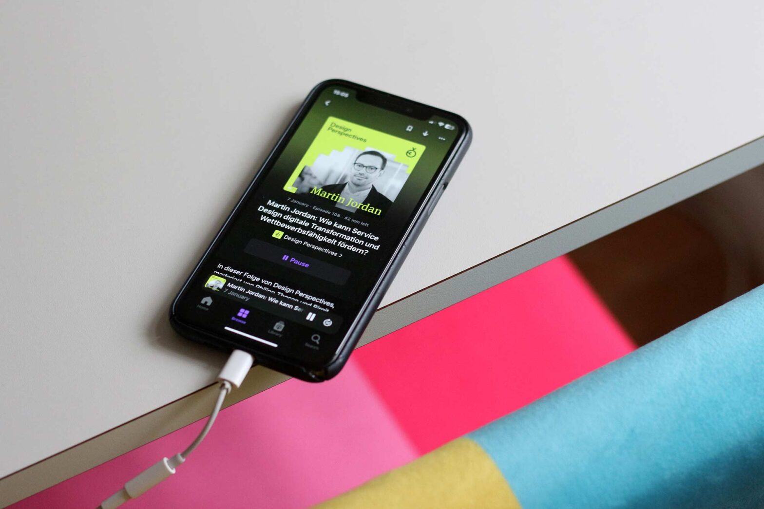 A phone showing a podcast playing on a grey desk with a colour chair in front – the screen shows a man