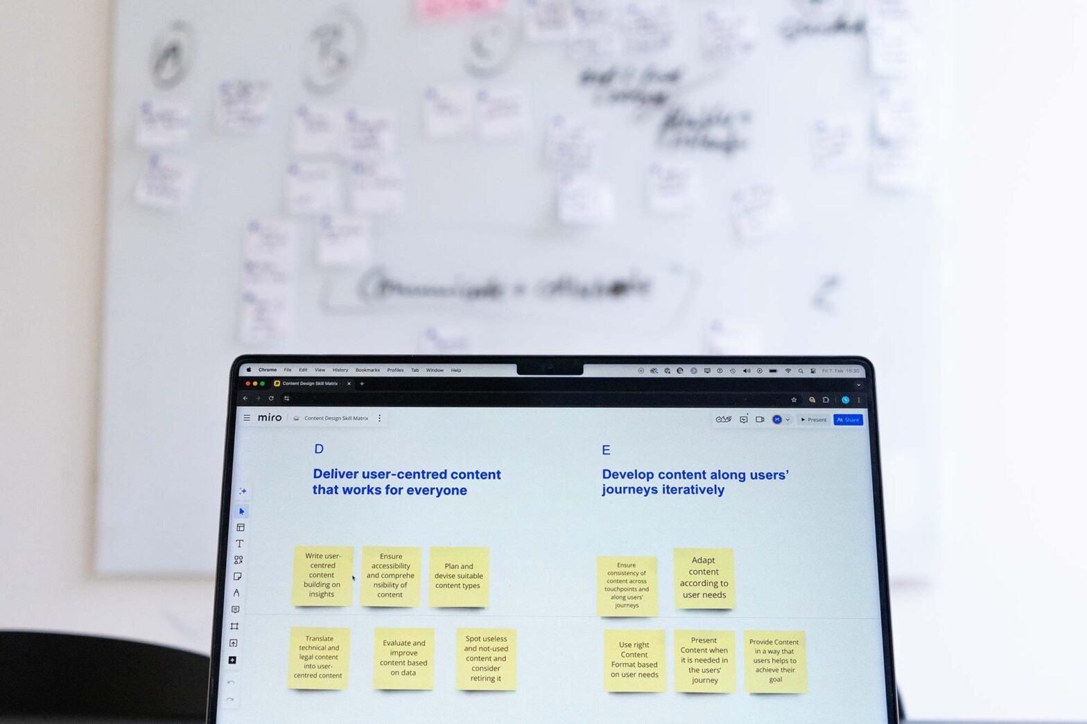 A laptop screen with a virtual whiteboard with 11 sticky notes in front of a physical whiteboard with further clustered notes