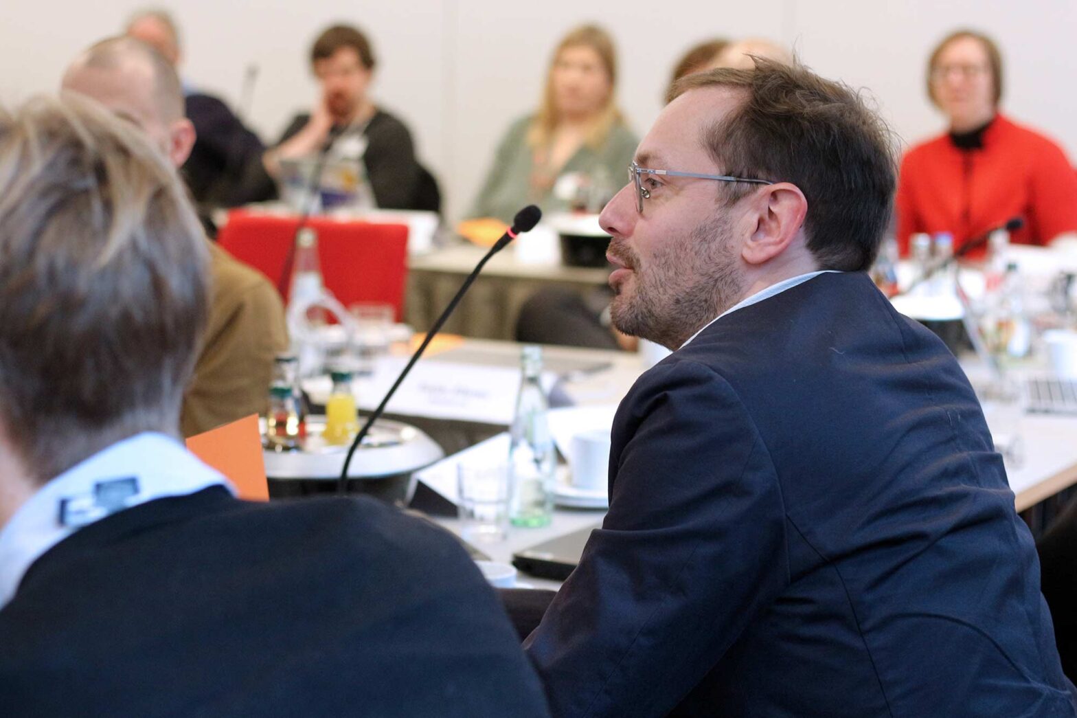 A man in a dark suit and with glasses speaking into a microphone during a meeting or conference. Other attendees are seated at tables in the background, with documents, water bottles, and coffee cups visible.