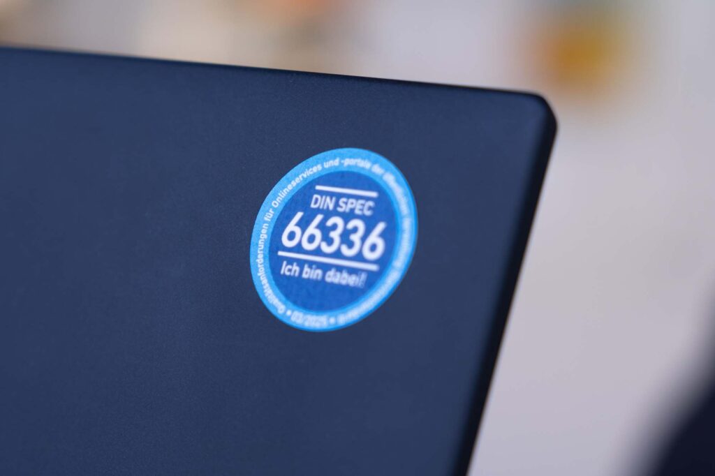 A sticker on a laptop saying: “DIN SPEC 66336 – I’m onboard!”