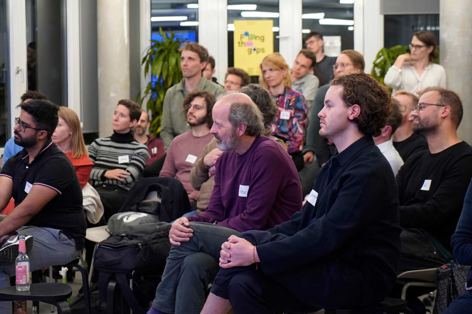 A group of about 20 people sitting in a modern office space paying attention to something outside of the picture, behind them at a window is a poster saying ‘Filling the gaps’