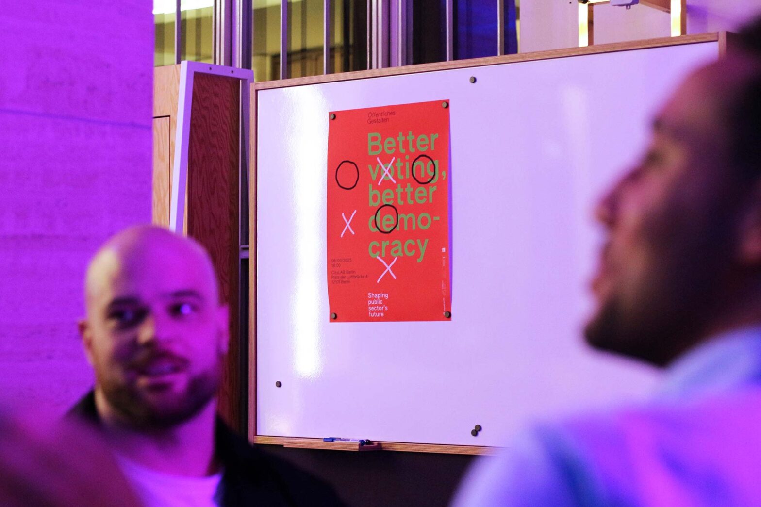 A red and green poster with the text "Better voting, better democracy" is pinned to a whiteboard in a room with purple lighting. Two people in the foreground are engaged in conversation, slightly blurred. The setting appears to be an event or discussion on civic engagement.