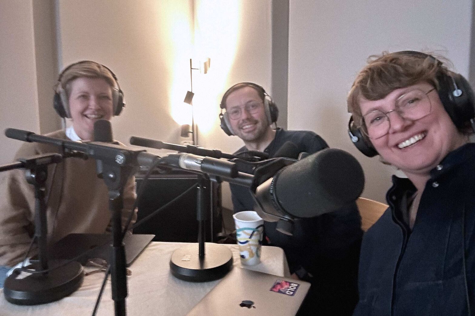 Selfie of a woman with light skin together with 2 people – another woman and a man; all 3 sit in front of professional microphones, wear big headphones and smile into the camera