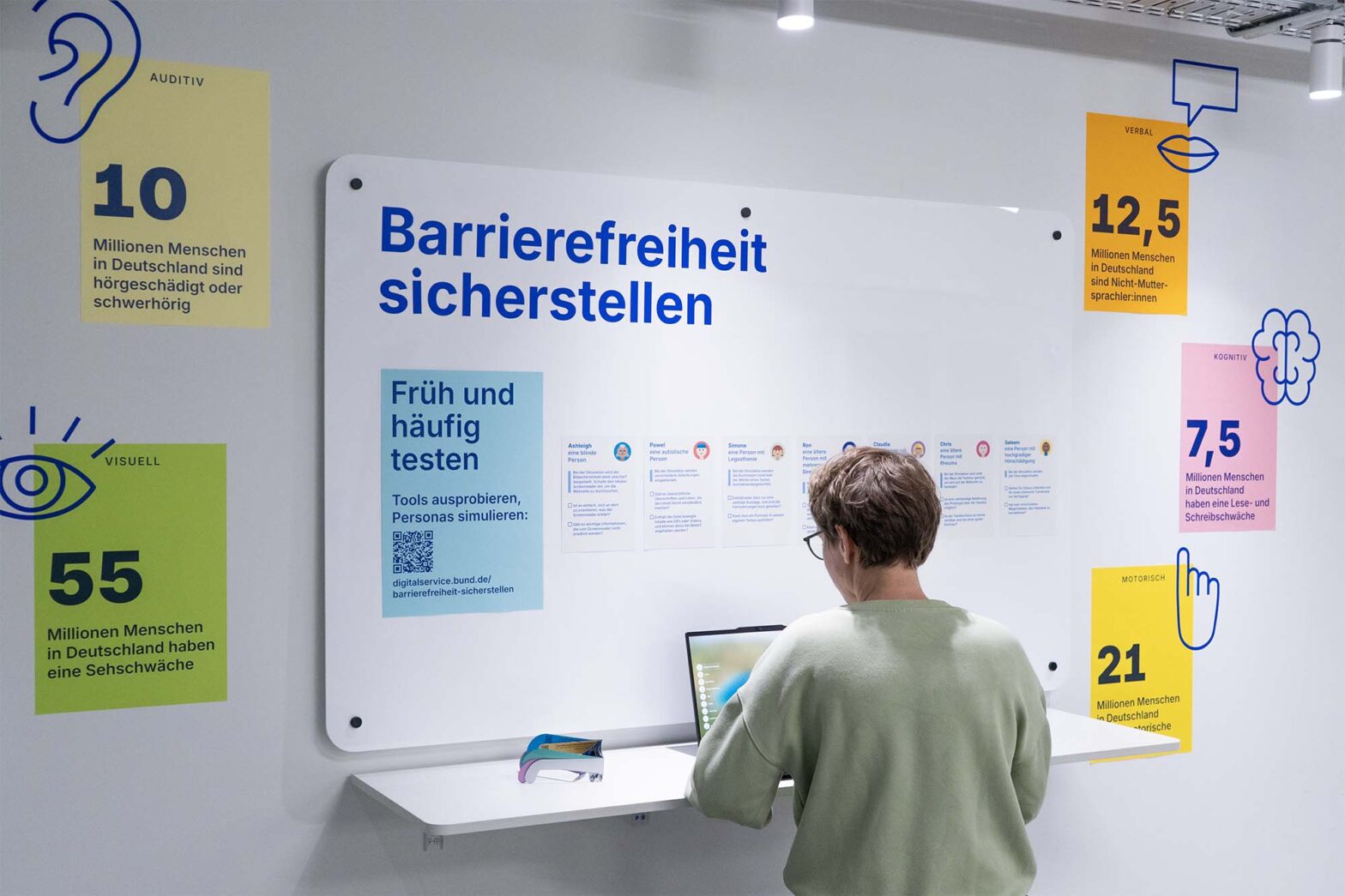 A person sits at a white counter engaging with an interactive display about accessibility. Surrounding infographics highlight statistics on disabilities in Germany, including visual, auditory, motor, cognitive, and verbal impairments.