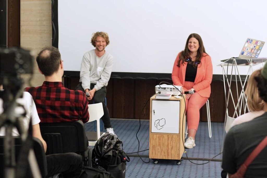 2 people, 1 man and 1 woman, both with light skin sitting on chairs in front of an audience holding microphones; both wear semi-casual cloth and are smiling
