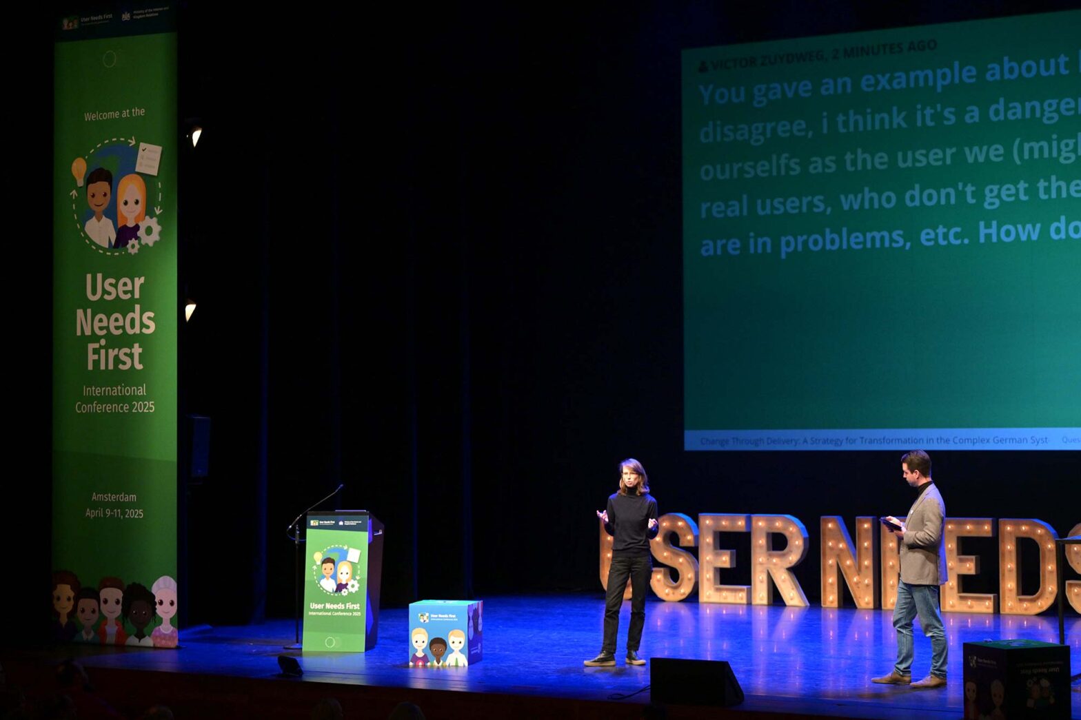 A woman in an all-black casual outfit is presenting on a big stage with a lectern and banner next to her; a moderator is right of her with ample light bulbed letters saying “User Needs“ behind them on the stage