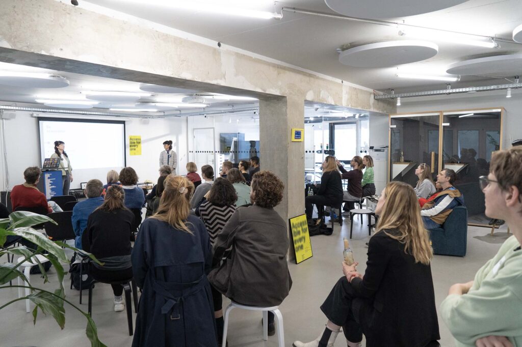 A group of some 30 to 40 people stting in a modern office space listening to 2 women standing in front of a projected canvas and next to a lectern; a poster leaning against a column says ‘accessible for everyone’