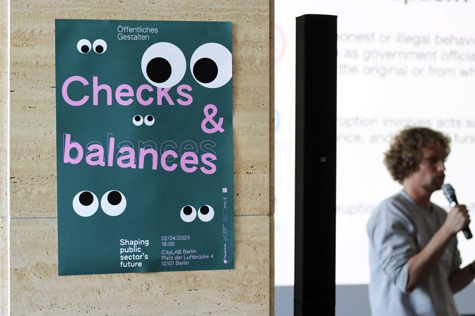 Poster on a marble column with several pairs of googly eyes in different sizes saying: “Checks & balances” in big letters – with the word balances hanging down; in the background, a person with curly hair is speaking with a microphone