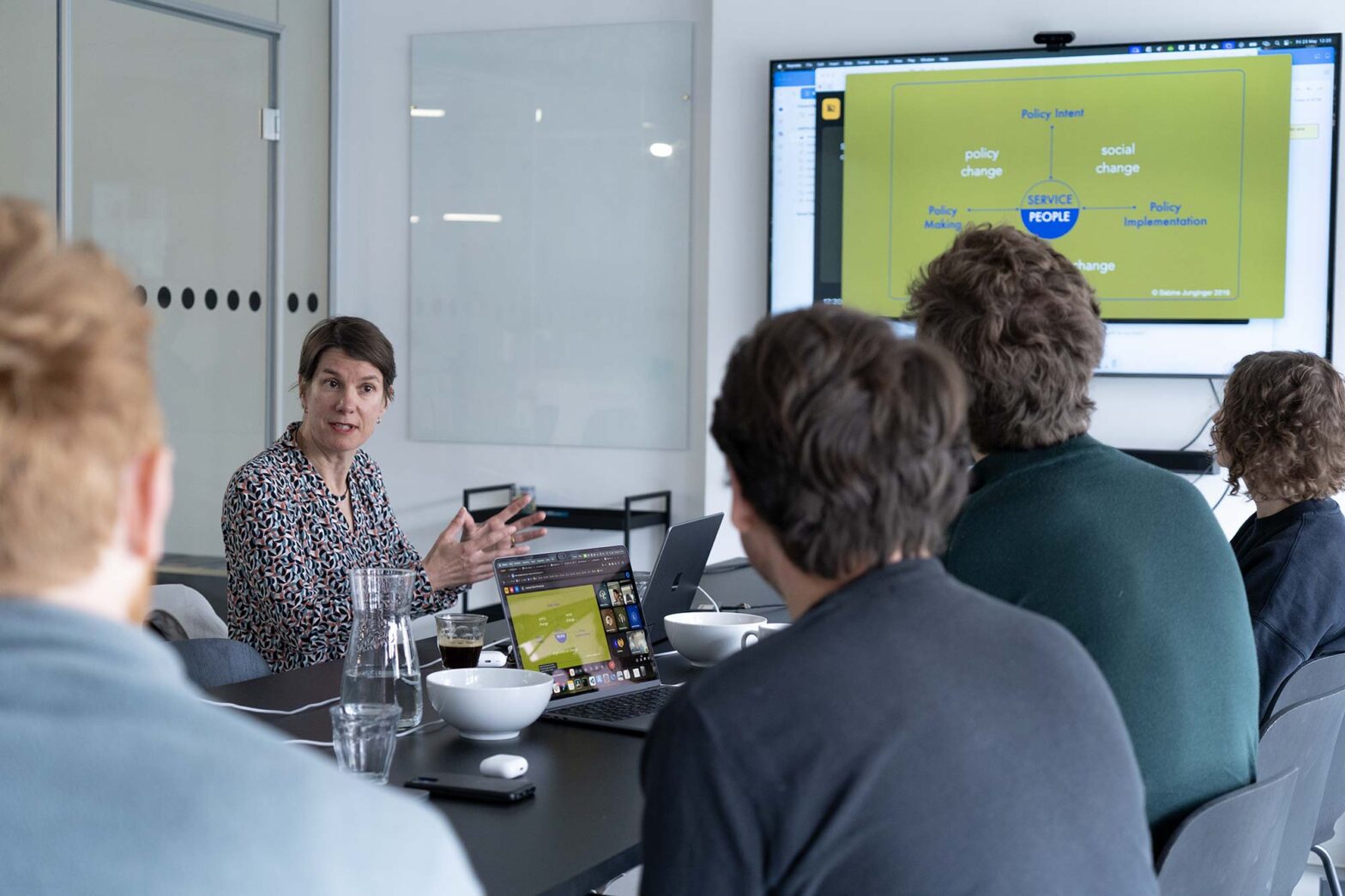 A woman leads a discussion with a group of people in a conference room, with a presentation slide about policy change and service design displayed on a large screen.