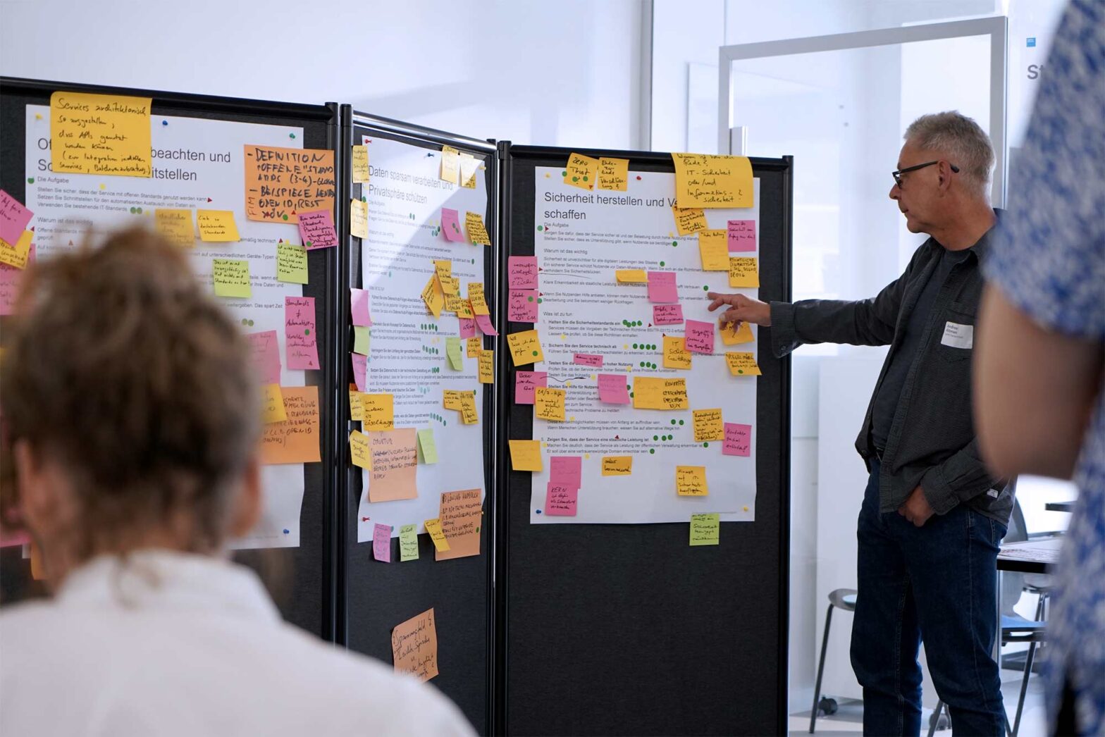 A man with grey hair standing at a 3-part wall with posters covered in coloured sticky notes, pointing and talking, 2 other people in the foreground are looking at him.