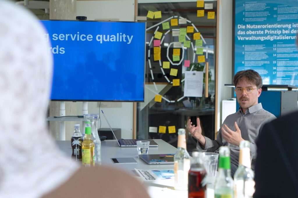 Several people are sitting at a conference table with bottles of water and notebooks on it. A presentation with the partially visible inscription “service quality” is running on a screen. One person is talking and gesticulating with their hands. In the background, notes are stuck to a glass wall. Next to it is a poster with the inscription “User orientation is the overriding principle of administrative digitization.”