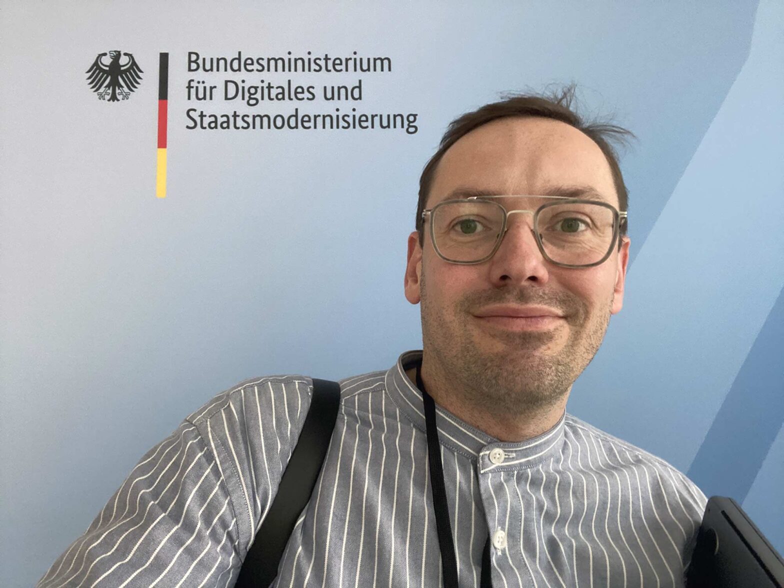 Selfie of a light-skinned middle aged man wearing a lined blue shirt in front of a light blue wall with the logo of the Federal Ministry for Digital and State Modernisation