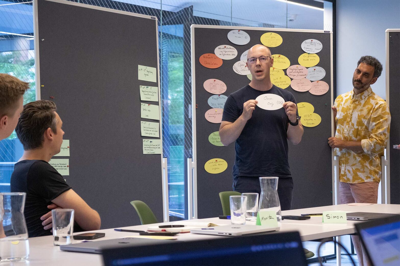 A man stands in front of a group during a workshop, holding a white oval card with the words “Once - Only (OO)” written on it. Behind him, colorful sticky notes are pinned to a board. Three other men are present, two seated and one standing, listening attentively. The setting appears to be a modern office or conference room.