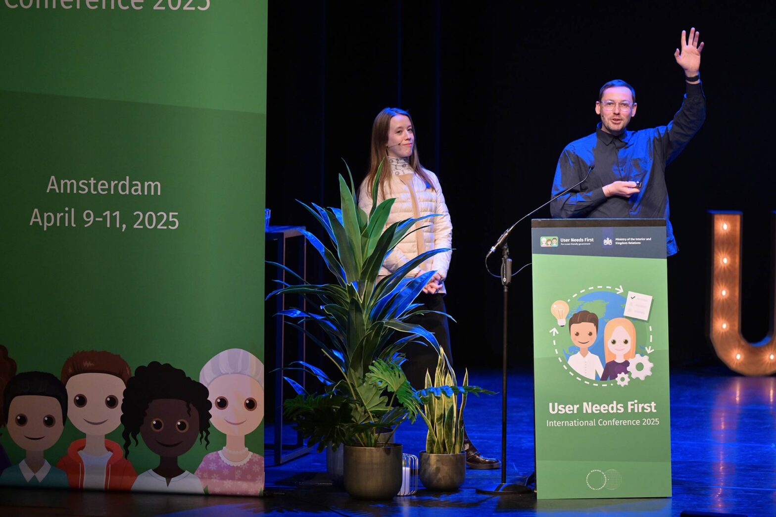 Two speakers stand on stage at the “User Needs First” International Conference 2025 in Amsterdam, with one speaking passionately at a lectern.