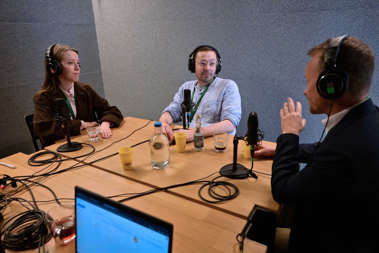 Three people, one woman and two men, sitting around a wooden table in a small, soundproofed space, all wearing big headphones and having professional microphones in front of them