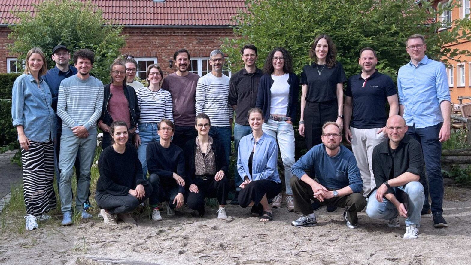 Group picture of 19 people standing together, smiling in front of a refurbished farm house