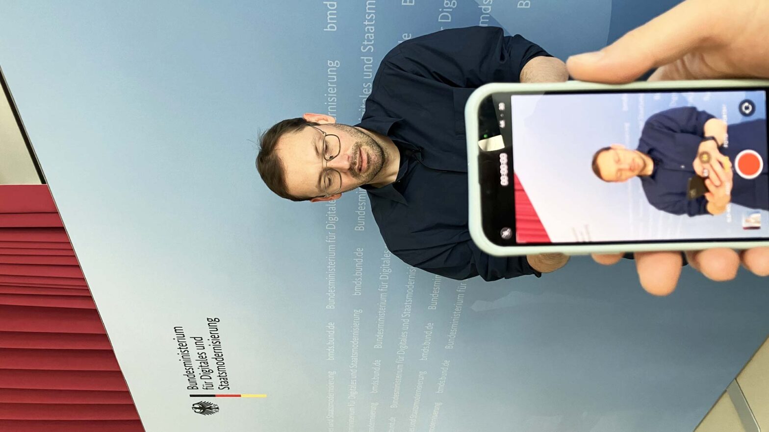 A light-skinned man in front of a presentation wall with the logo of the Federal Ministry for Digitalisation and State Modernisation; a phone is in front of him recording him while he is also a holding a phone