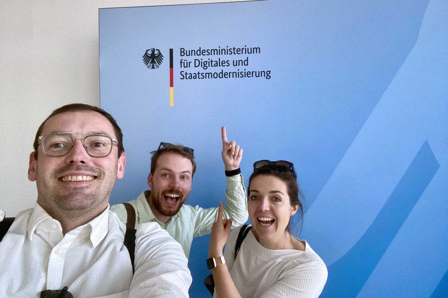 Selfie of 3 light-skinned people, 1 woman and 2 men, in front of a light blue wall with the logo of the Federal Ministry for Digital and State Modernisation – all smiling, 2 of them pointing at the logo