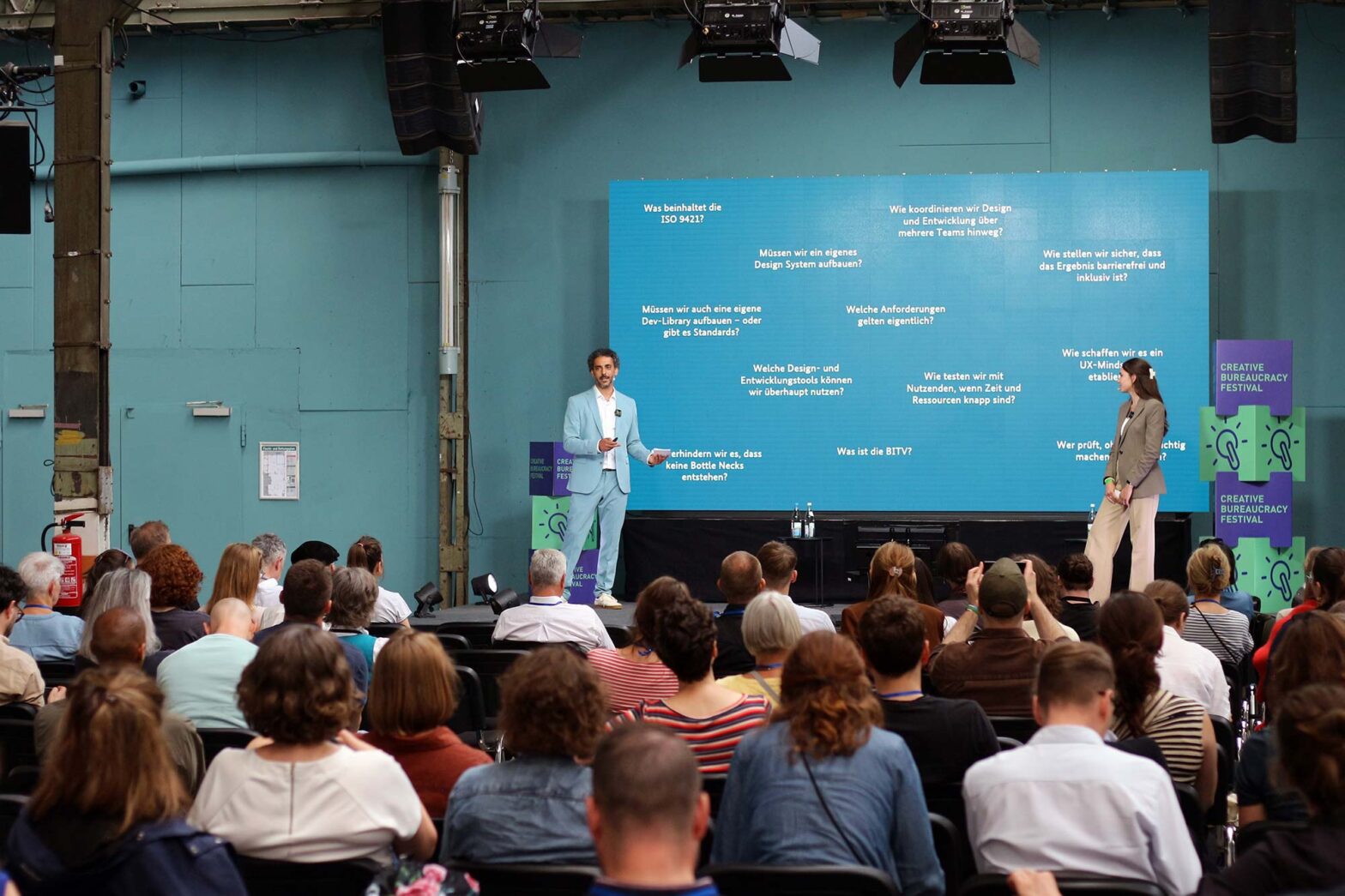 A man and a woman are speaking on stage at the Creative Bureaucracy Festival, with a screen behind them displaying questions in German to an audience seated in front.