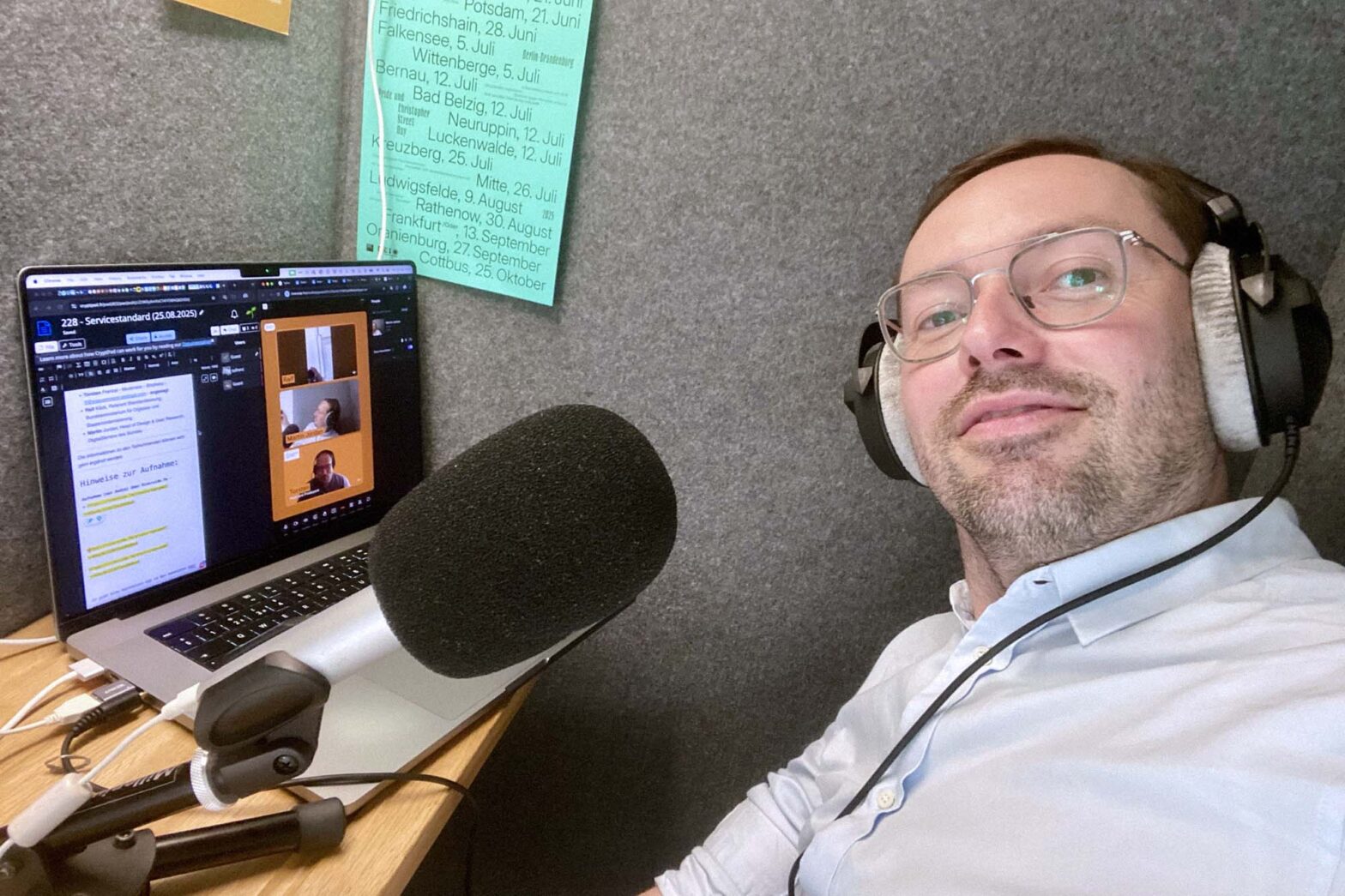 Selfie of a light-skinned man with glasses, wearing a light blue shirt and huge over-ear headphones, sitting in a felt-covered cabin with a huge microphone and a computer showing notes and other people dialled in with video