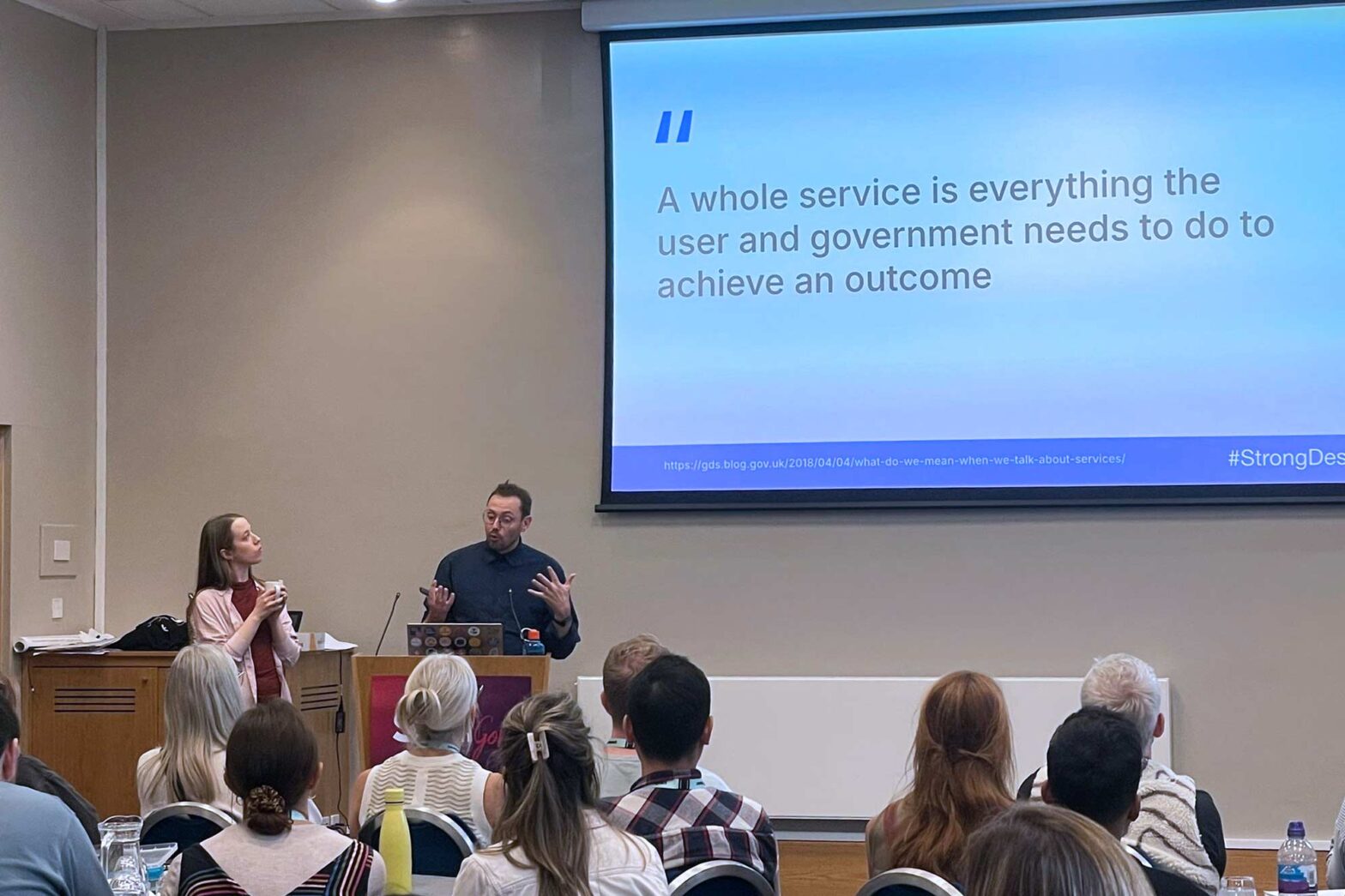 2 light-skinned people, a man and a woman in a conference room speaking in front of an audience; a projected slide above the gesticulating man in front of a lectern contains a quote: “A whole service is everything the user and government needs to do to achieve an outcome”