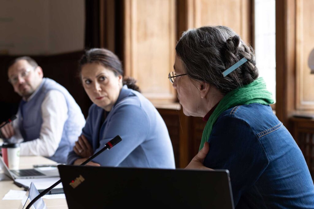 3 people in a conversation, 2 middle-aged light-skinned women and a man – all with conference space table microphones and laptop computers in front of them, 1 woman is in focus, she seems to be speaking while the other 2 people are looking at her, listening