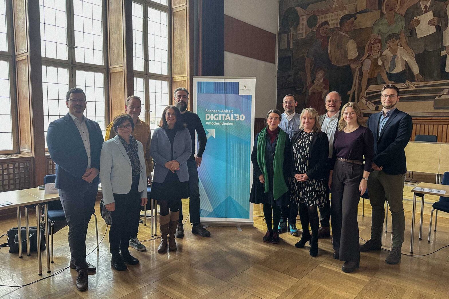 A group of middle-aged white people, 5 women and 6 men, standing next to a roll-up banner that says ‘Digital 30’ in a modernist 20-century town hall room with a workers movement mural in the back