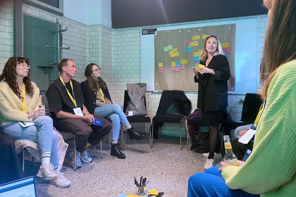 A workshop setup in a converted space with 5 people sitting in a semi-circle and a woman standing in front of a white board; she is holding a pen and sticky note pad, about 2 dozen stickies notes are on the wall behind her; everyone is wearing yellow lanyards, indicating they attend some event