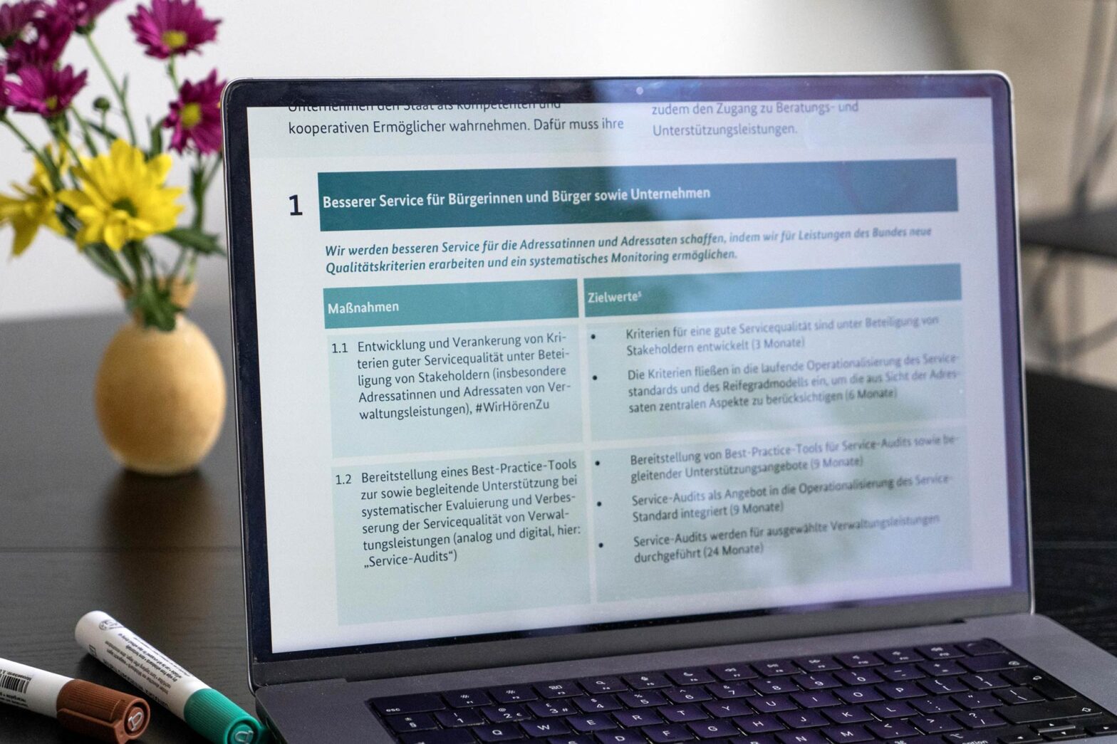 A laptop is on a dark table, displaying a presentation slide with German text. A white vase with purple and yellow flowers is to the left of the laptop, and two marker pens are at the bottom left. The keyboard is visible in the foreground