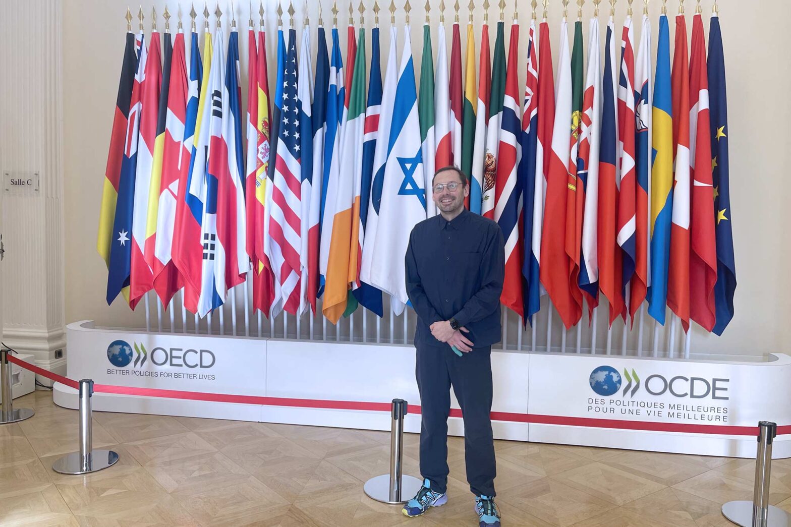 A middle-aged man standing in a official setting in front of a row of dozens of flags; with logos of the OECD below – and the tagline ‘better policies for better lives’; the man is looking into the camera and smiling