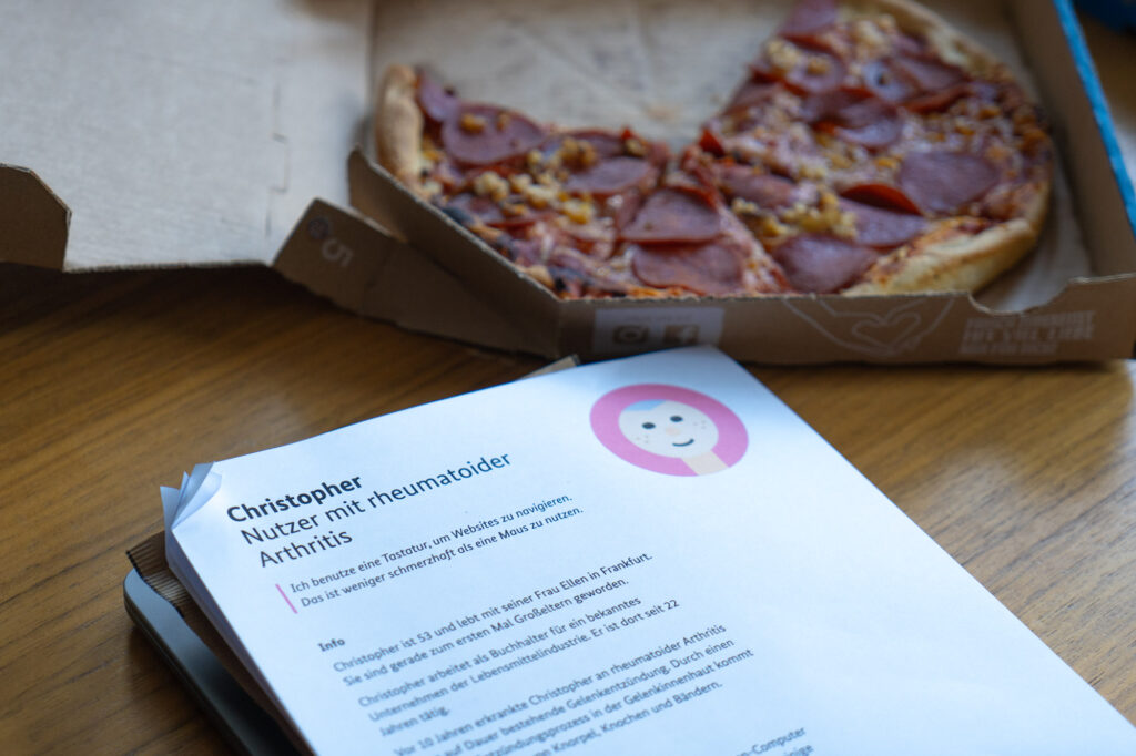 A pile of papers with the description of an accessibility persona named Christopher, a user with rheumatoid arthritis, on a table next to a 3/4 salami pizza