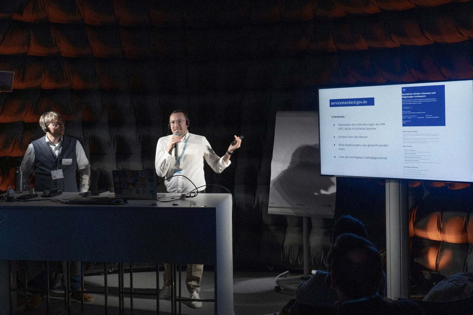 A presentation is being given inside a large, inflatable dome. 2 men are at the front, one seated and one standing at a podium holding a microphone. On the right, a large screen displays a presentation slide with German text.
