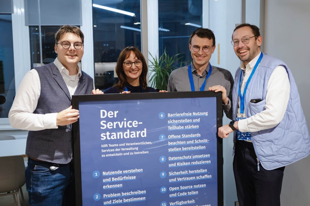 4 light-skinned people, 1 woman and 3 men posing with a framed poster of the Service Standard listing its criteria in a modern office space in front of a glass wall – all are smiling into the camera