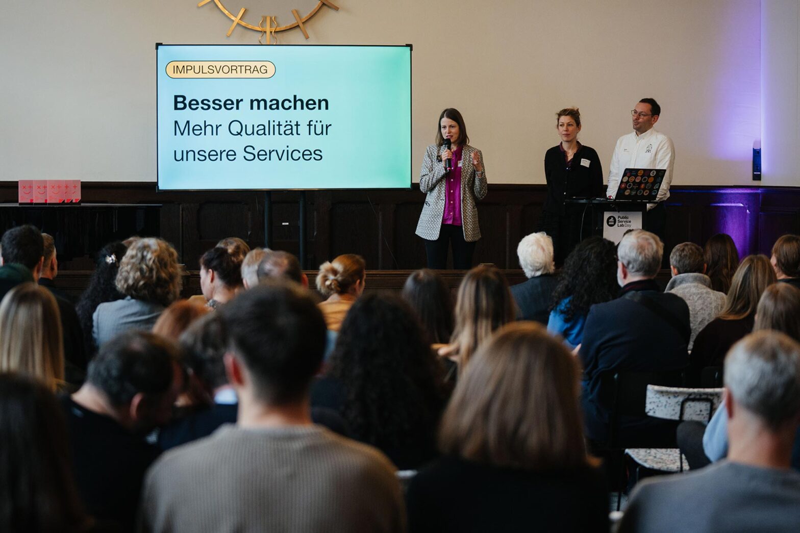 3 Personen, 2 Frauen und 1 Mann sprechen vor einer größeren Gruppe Menschen bei einer Veranstaltung; neben ihnen steht ein großer Fernseher, darauf steht: ›Impulsvortrag: Besser machen – Mehr Qualität für unsere Services‹