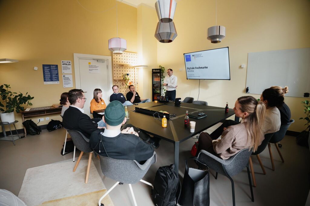 
A yellow-painted room with a large central table setup, 9 people are sitting around the table in O-shape, pens and sticky notes suggest a workshop setup, a man in a white shirt is standing in front of a mounted television, which shows a screen saying ‘digital umbrella brand – workshop‘ with the logos of the DigitalService and the Federal Ministry for Digitalisation and Government Reform